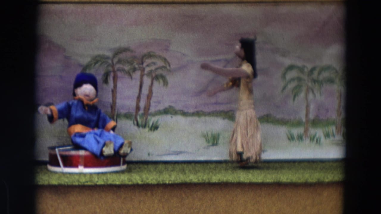 Blue-clad puppet seated on a red drum beside a grass-skirt dancer figure on a small stage.