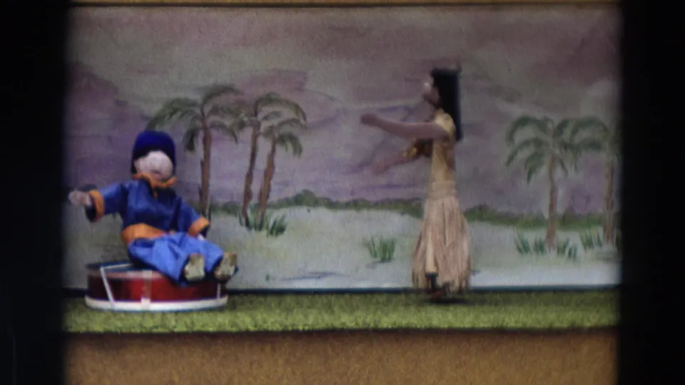 1968 Lahaina Maui 8mm frame showing a blue puppet on a red drum and a grass-skirt figure before a painted palm-tree backdrop.