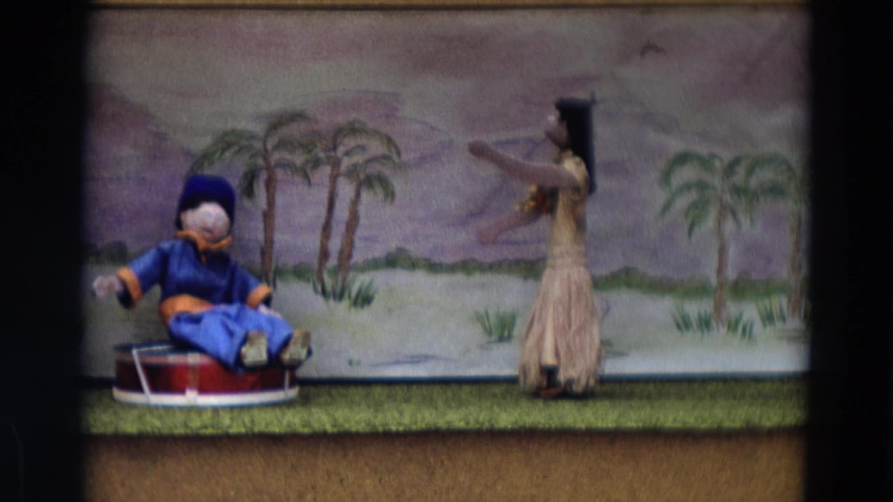 1968 Lahaina Maui home-movie frame of a puppet show, with a blue seated puppet and dancer figure before a tropical backdrop.
