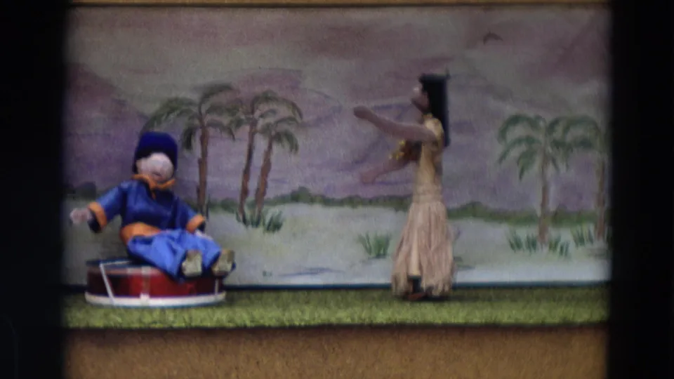 1968 Lahaina Maui home-movie frame of a puppet show, with a blue seated puppet and dancer figure before a tropical backdrop.