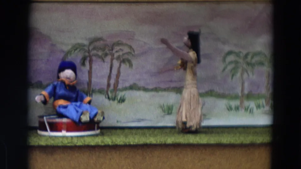 1968 Lahaina Maui 8mm frame with a grass-skirt dancer figure raising an arm near a blue puppet on a red drum.