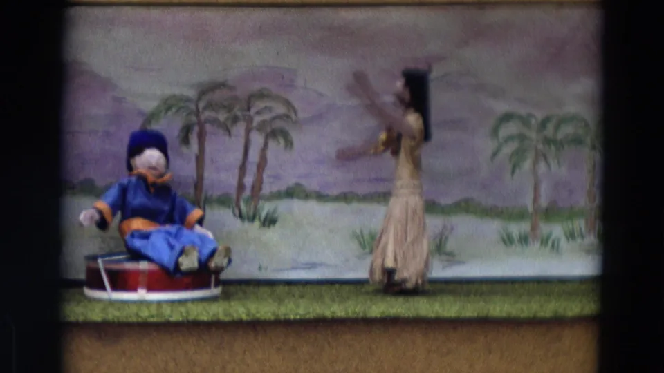 Final 1968 Lahaina Maui frame showing the puppet-stage arrangement with blue seated puppet, dancer figure, palms, and painted hills.
