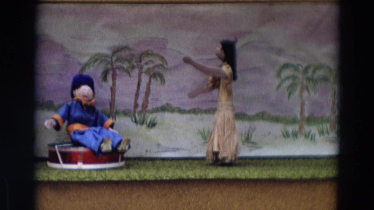 Seated blue puppet on a red platform beside a standing skirted puppet on a small stage.