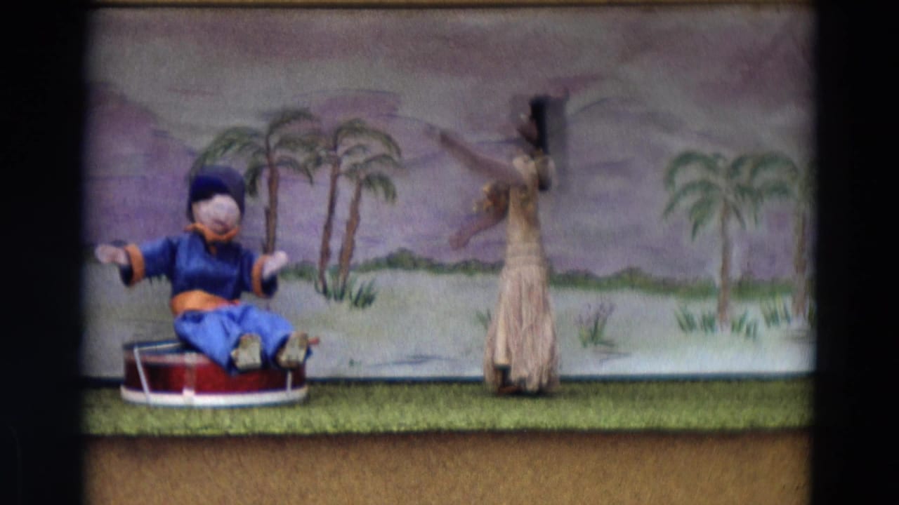 Blue seated puppet gestures while a standing skirted puppet faces the painted palm backdrop.