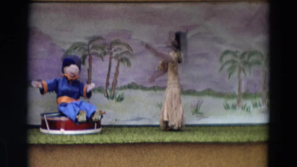 1968 Lahaina Maui 8mm frame with a blue seated puppet gesturing near a standing skirted puppet and tropical painted scenery.