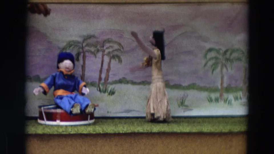 1968 Lahaina Maui 8mm frame with a standing grass-skirted puppet gesturing beside a seated blue puppet in a painted tropical set.