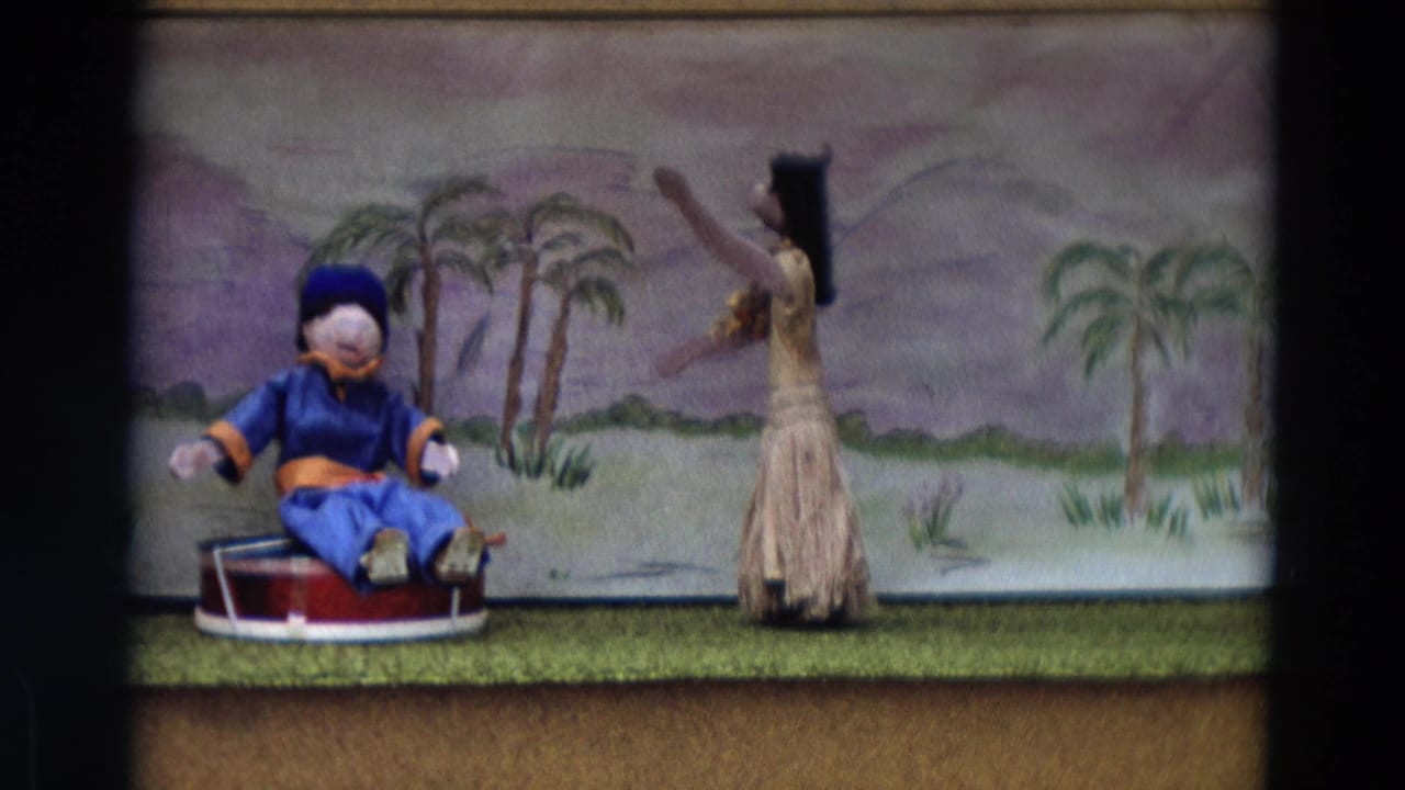 Male puppet in blue sits on a red drum while a female puppet dances before a palm-tree backdrop.