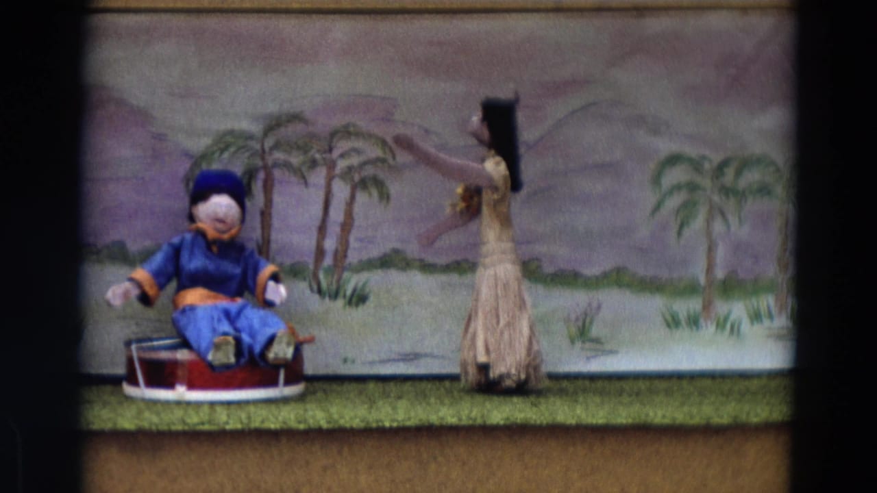 Two puppets appear on a small stage with painted palm trees and hills behind them.