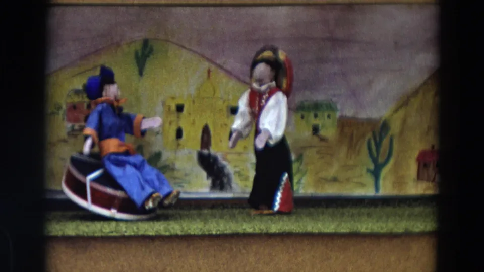 1968 Lahaina, Maui frame showing a male puppet on a red drum facing a female puppet in front of a painted village backdrop.