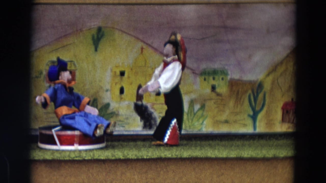 1968 Lahaina Maui home-movie still of two handmade puppets staged against painted hills, cacti, and adobe-style buildings.