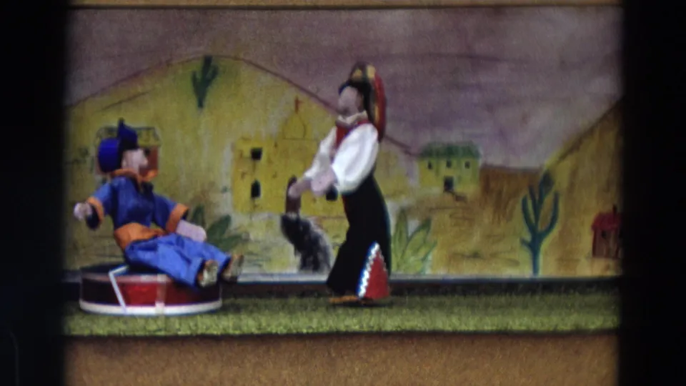 1968 Lahaina Maui 8mm still showing a blue-clad puppet seated low as a second costumed puppet stands near a painted desert backdrop.