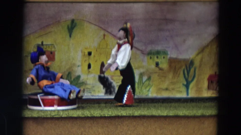 1968 Lahaina Maui 8mm frame showing a blue-costumed puppet on a red drum facing a standing puppet before a Mexican village backdrop.