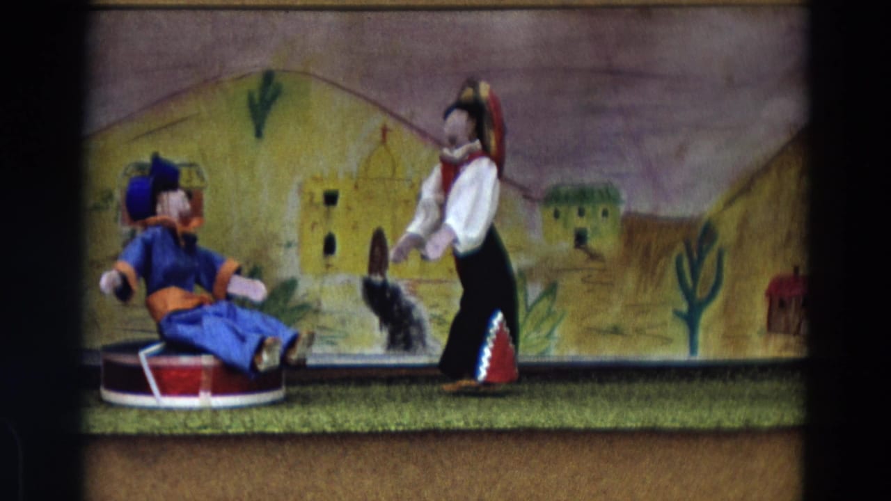 1968 Lahaina Maui 8mm frame of two handcrafted puppets posed in front of adobe-like painted buildings and desert plants.