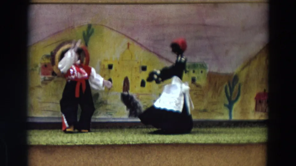 1968 Lahaina Maui 8mm frame with two puppets in Mexican-themed costume before painted adobe-style buildings and cacti.