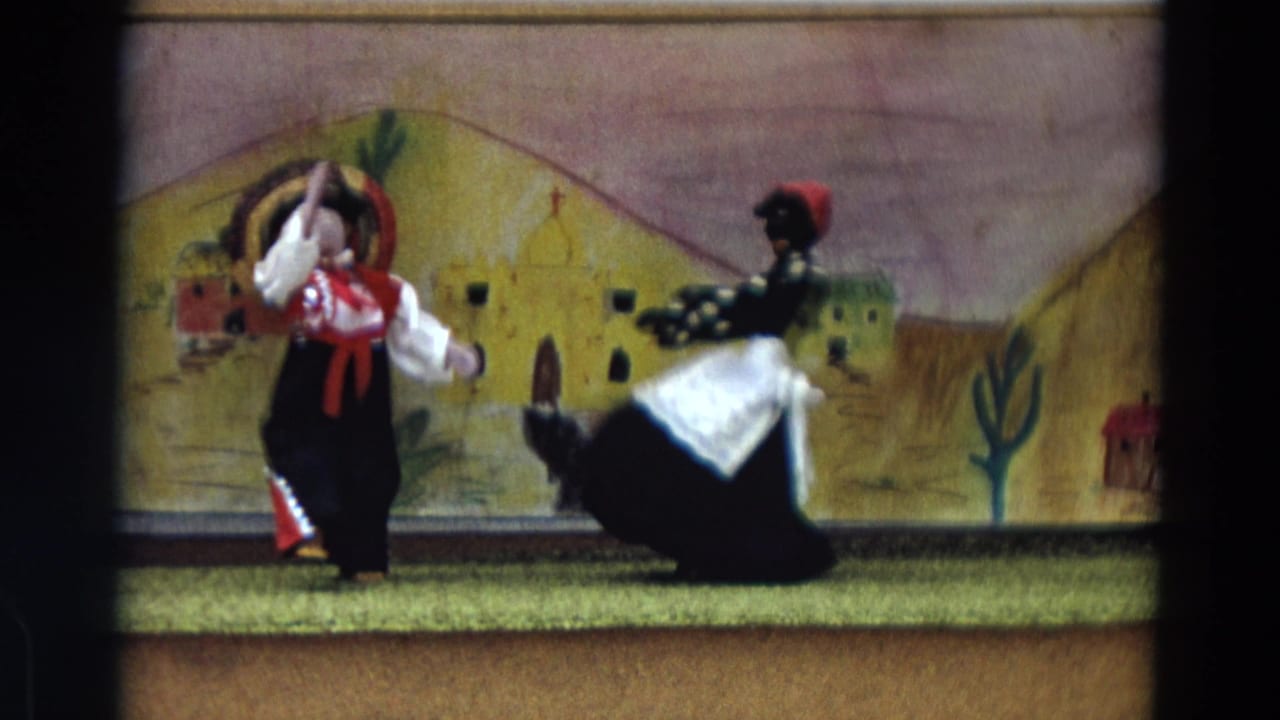 The female doll in a black dress and white apron turns toward the male doll against the painted Lahaina performance backdrop.