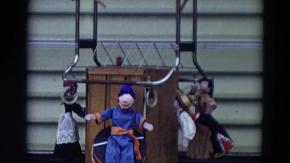 1968 Lahaina, Maui frame showing a simple puppet stage with a blue-costumed puppet, hanging figures, metal rods, and a wooden box backdrop.