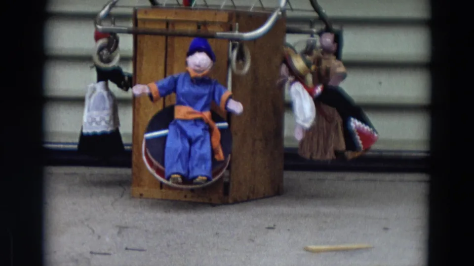 1968 Lahaina, Maui home-movie frame showing a blue-clad puppet seated in a wooden box with other puppets hanging nearby.