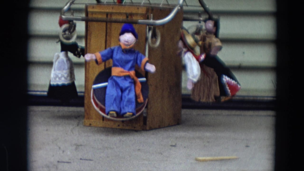 1968 Lahaina, Maui frame with a seated blue puppet in a wooden box and several hanging puppets beside it.