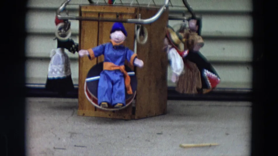 1968 Lahaina, Maui frame of a handmade puppet setup with a wooden box, metal bar, and several fabric figures.
