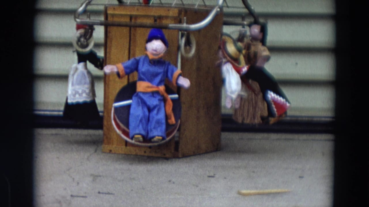Blue-clad seated puppet on a wooden frame with other hanging puppets nearby.