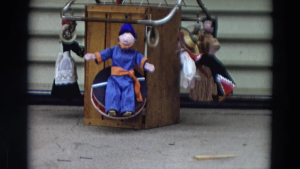 1968 Lahaina, Maui home-movie still with handmade puppets hanging around a wooden frame, centered on a blue seated figure.
