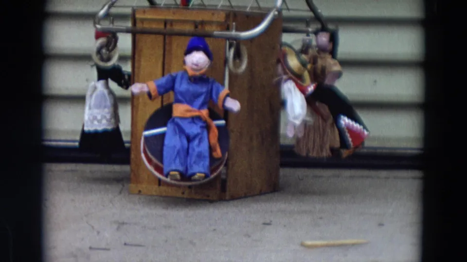 1968 Lahaina, Maui 8mm still showing the blue puppet slightly closer, with hanging figures and a pale wall behind it.