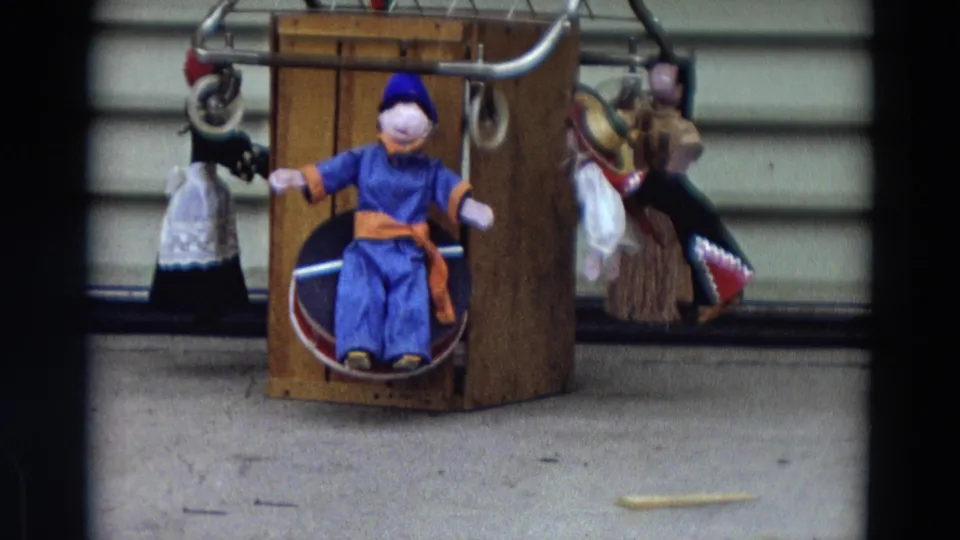 1968 Lahaina, Maui home-movie frame showing a blue seated puppet, orange sash, wooden frame, and background string puppets.