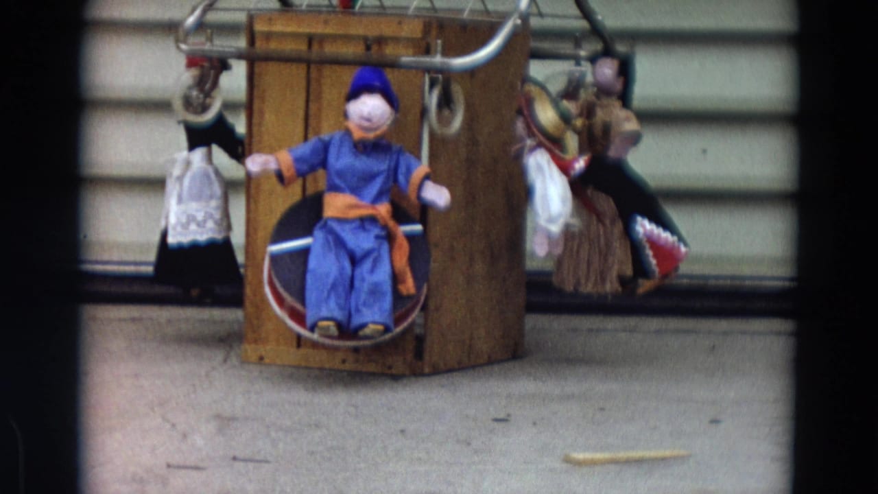 1968 Lahaina, Maui 8mm frame showing a blue-clothed seated doll with hanging dolls on a wooden display stand.