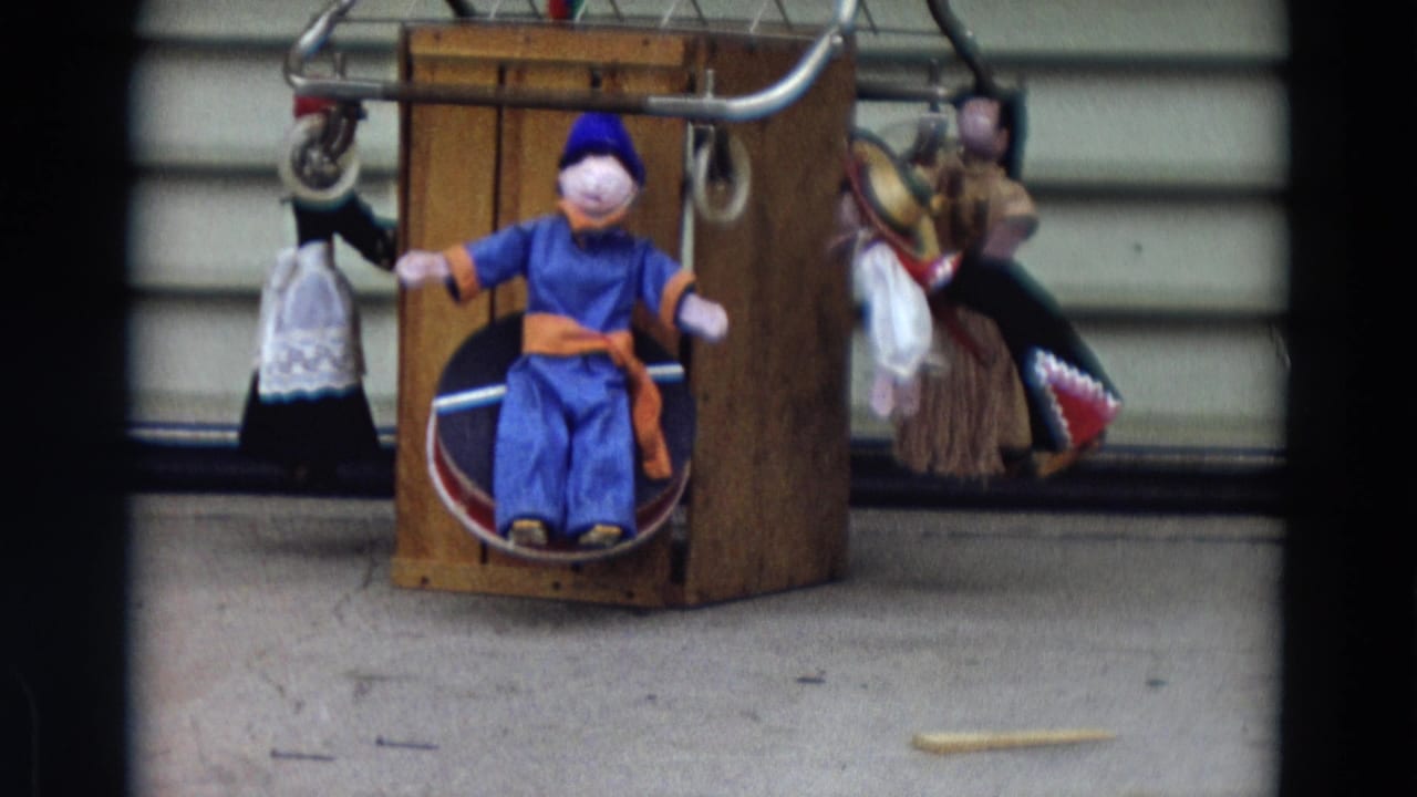 1968 Lahaina, Maui home-movie frame of a central blue doll on a wooden stand, flanked by smaller hanging dolls.