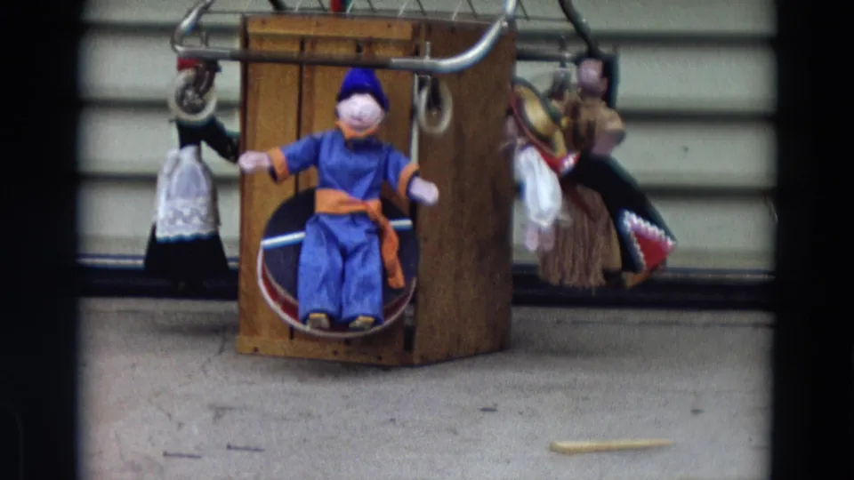1968 Lahaina, Maui home-movie frame of a central blue doll on a wooden stand, flanked by smaller hanging dolls.