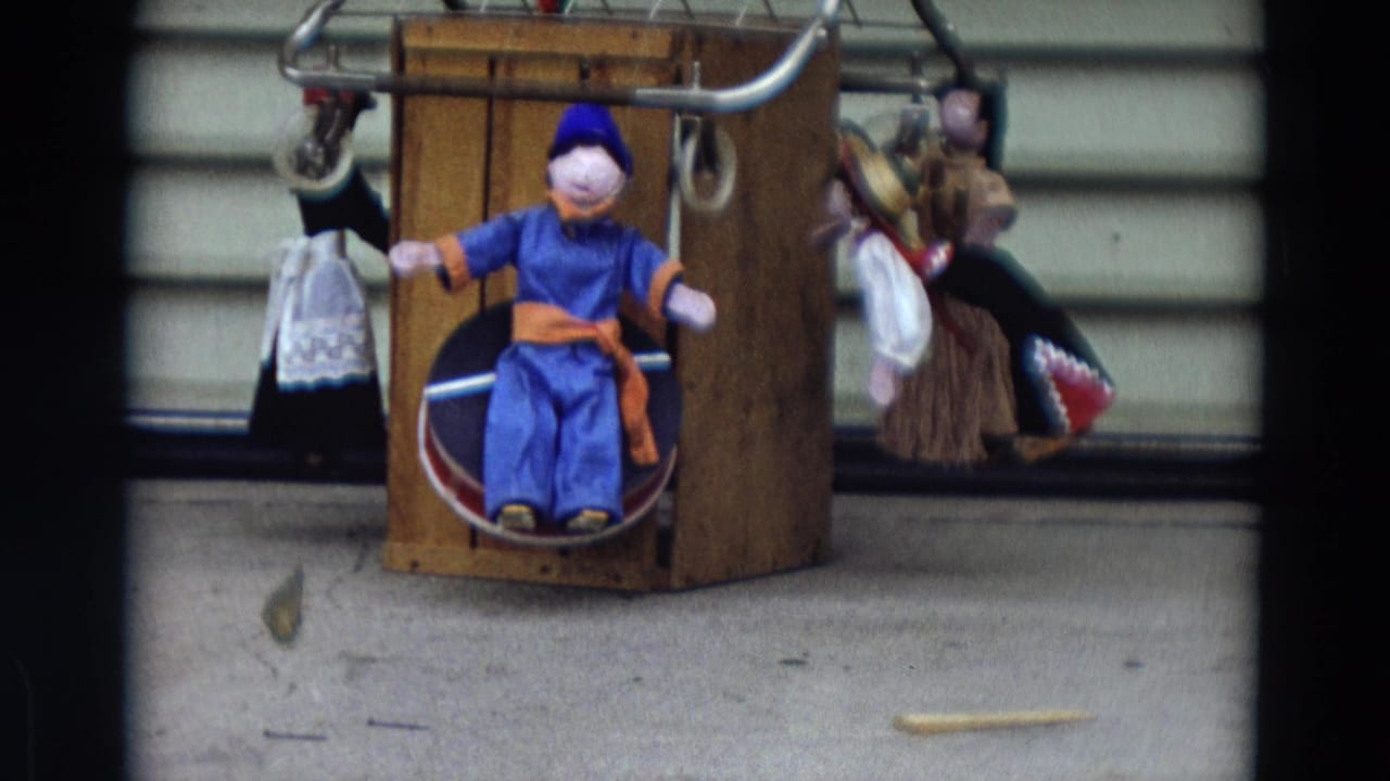 Blue-clad puppet seated in a wooden box with hanging puppets beside it.