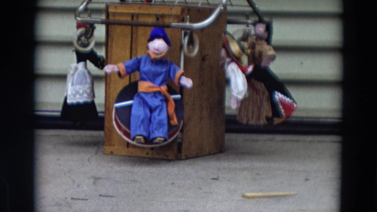 1968 Lahaina, Maui home-movie frame of a puppet display with a blue central figure, wooden stand, and hanging side figures.