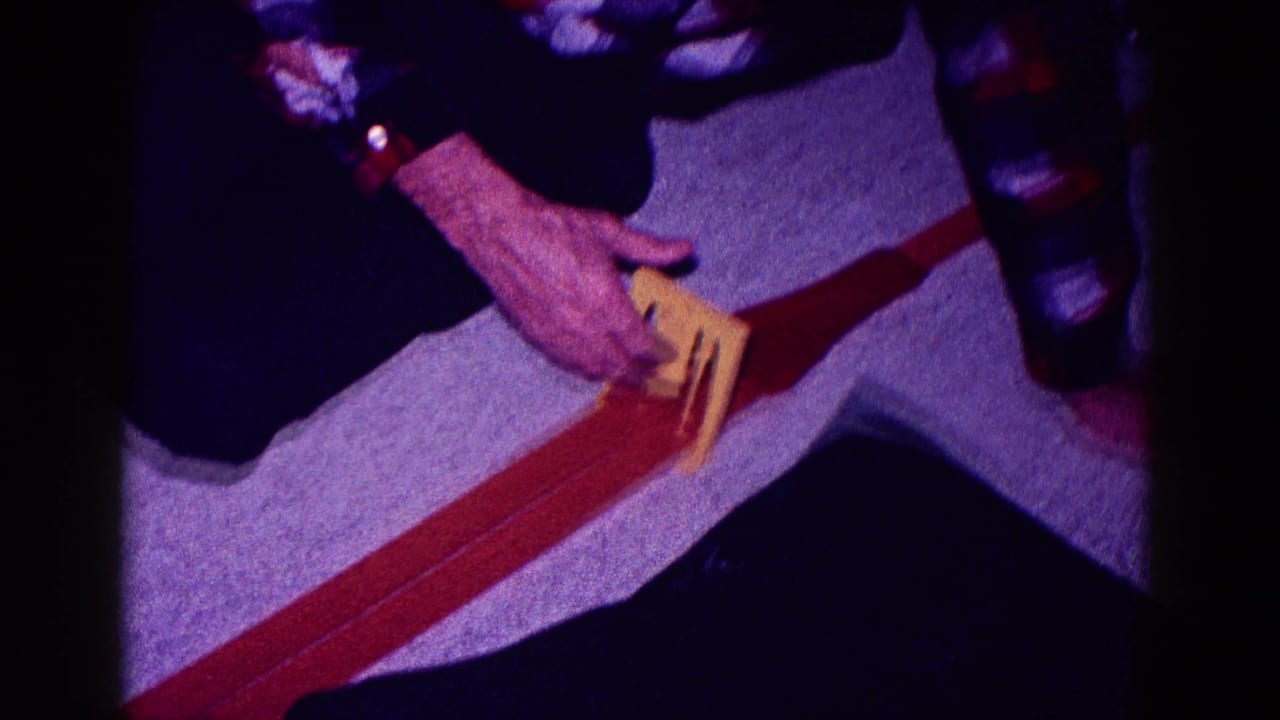 A man in a plaid shirt kneels indoors in 1968 Minnesota, positioning a yellow toy car on a red track.