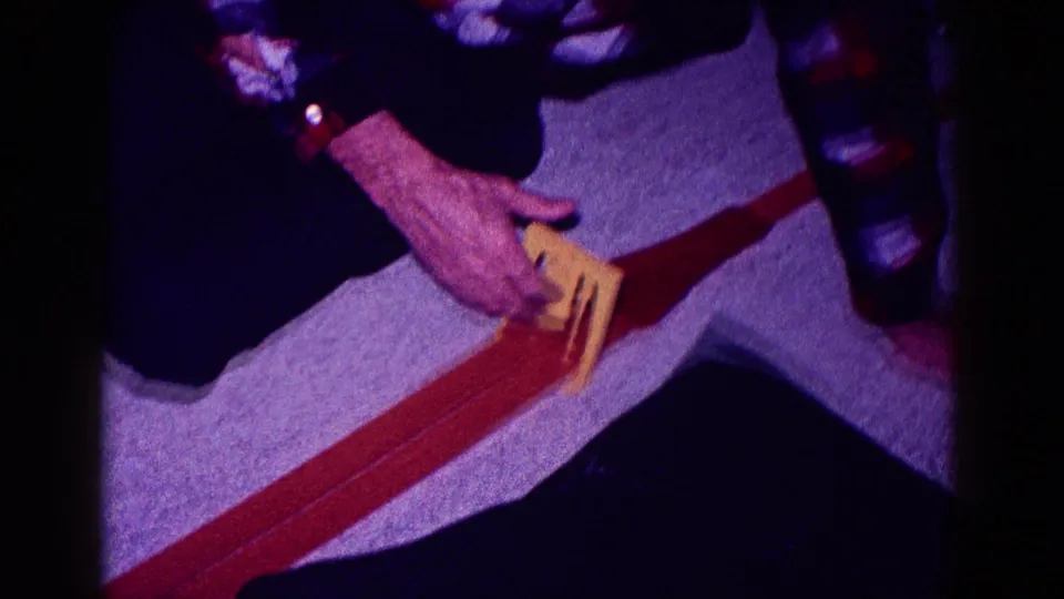 A man in a plaid shirt kneels indoors in 1968 Minnesota, positioning a yellow toy car on a red track.
