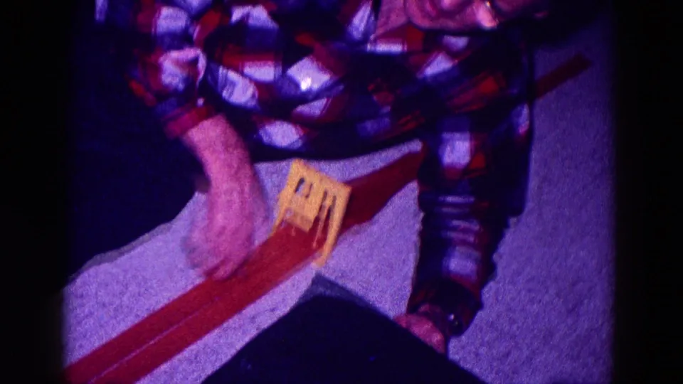 Overhead home-movie frame showing hands and a plaid sleeve as a yellow toy car is adjusted on a red track.
