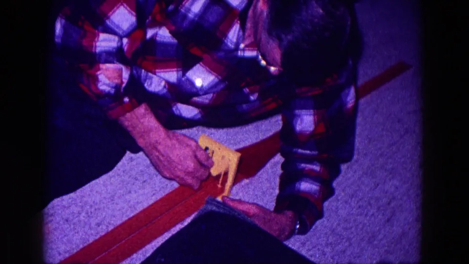 A kneeling man in a plaid shirt leans over a toy car track inside a Minnesota home-movie scene from 1968.