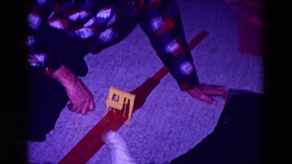 1968 Minnesota home-movie frame showing hands near fabric and a small wooden object on a carpeted private interior floor.
