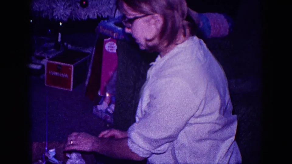 A woman in a light shirt bends over fabric work beside a red box in a dim private Minnesota interior from 1968.