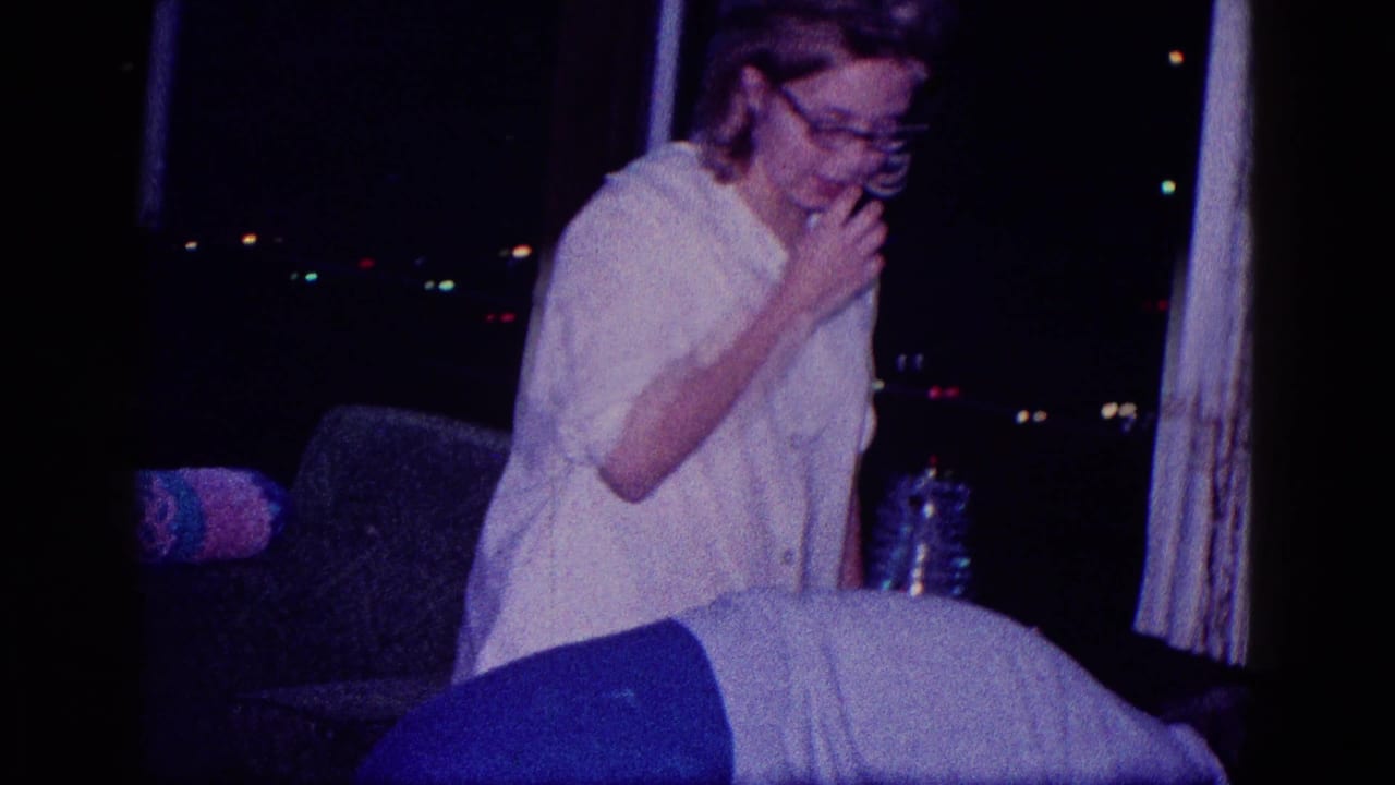 Woman in glasses sits or leans near a couch in a dim Christmas interior.
