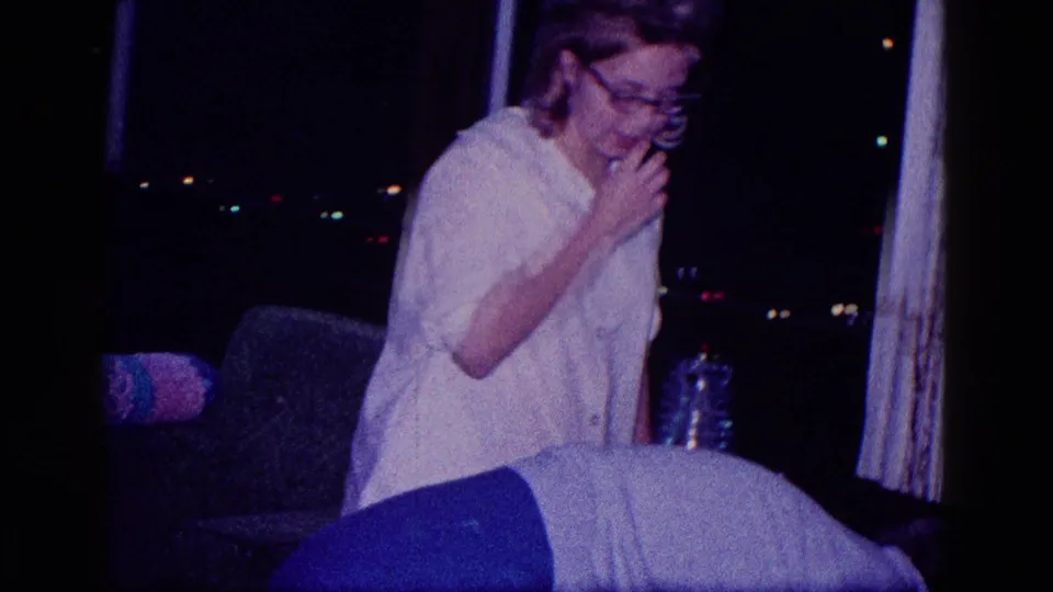 In a 1968 Minnesota home-movie frame, a woman in glasses and a white blouse sits or leans beside a couch in a dark holiday room.