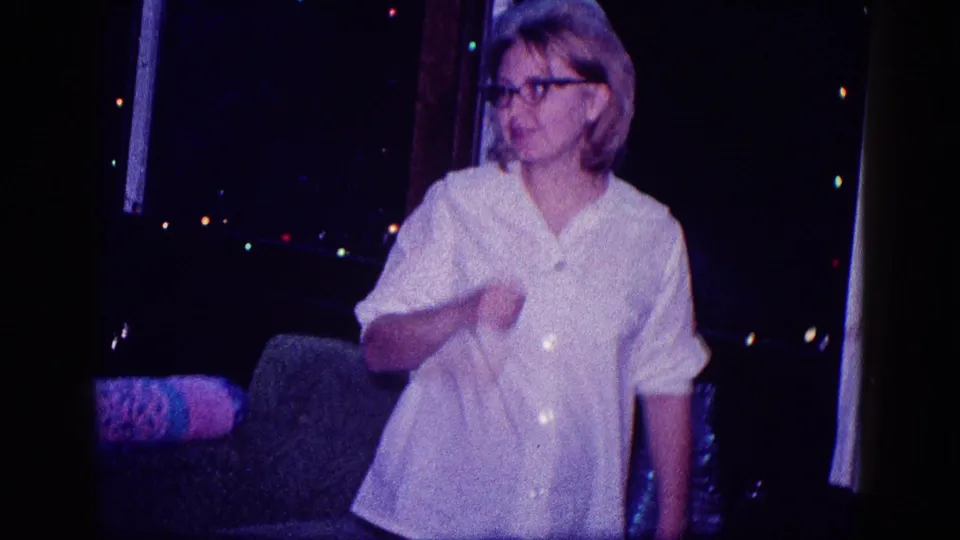 A woman in glasses and a white blouse stands in a dim Minnesota Christmas interior with a couch, curtains, and colored lights behind her.