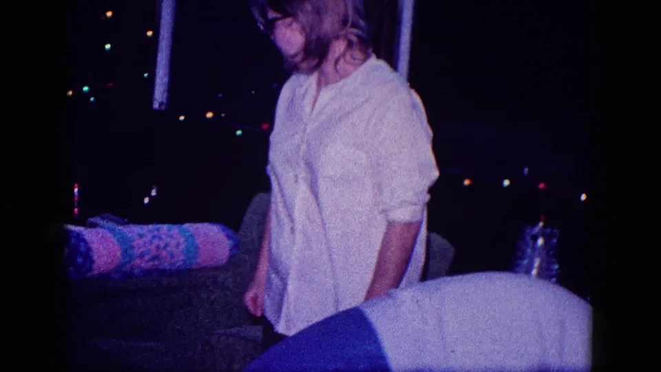 A woman in a white blouse moves past a couch inside a low-light 1968 Minnesota Christmas home-movie scene.