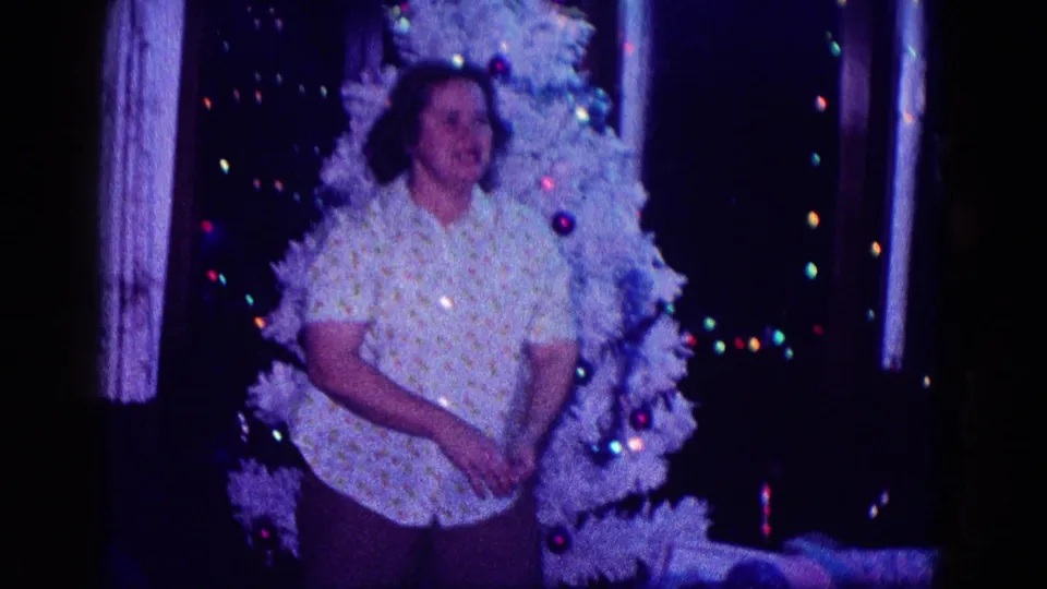 A woman stands before a white Christmas tree with ornaments and colored lights in a 1968 Minnesota home-movie holiday interior.