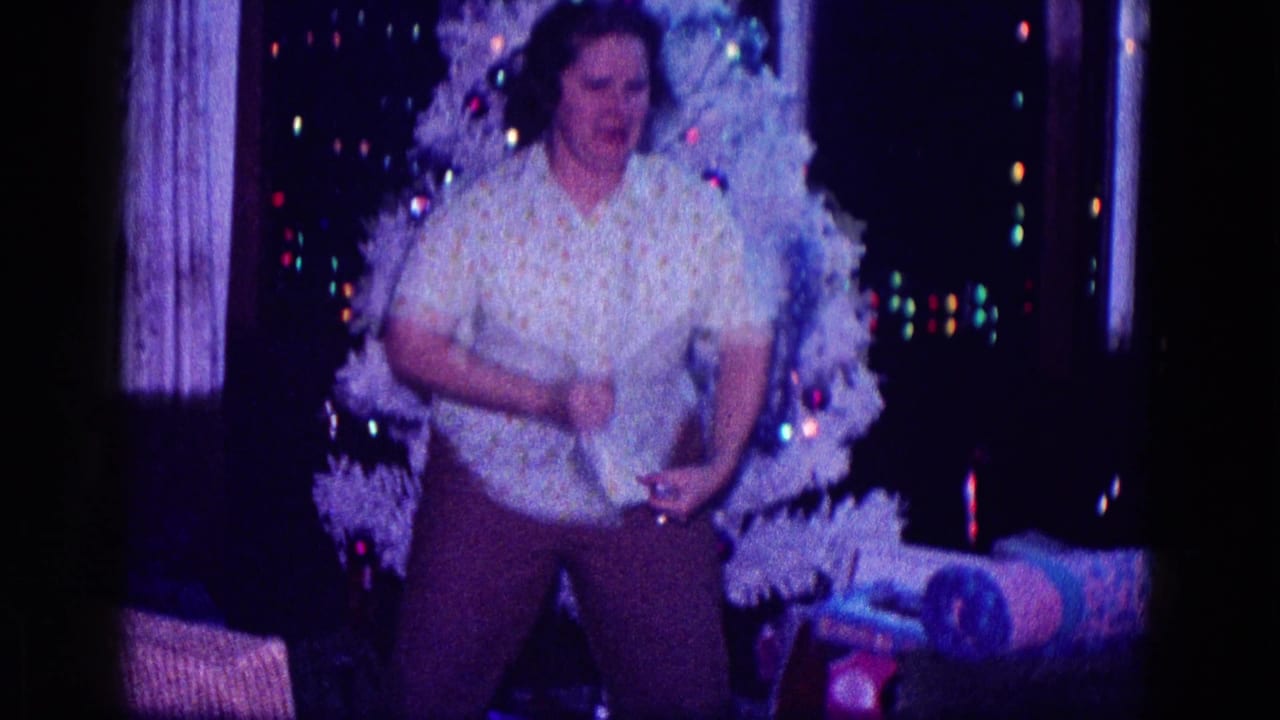 Woman gesturing in front of a lit white Christmas tree with gifts nearby.