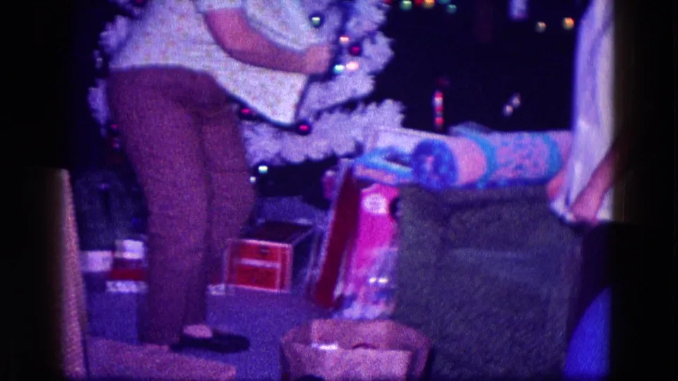 A cropped 1968 home-movie frame shows the woman from shoulders down beside the Christmas tree, with wrapped presents and furniture visible.