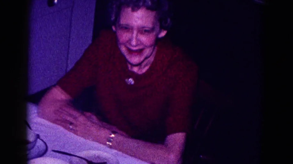 An older woman in a red sweater smiles toward the camera in the same dim private interior from the 1968 Minnesota holiday footage.