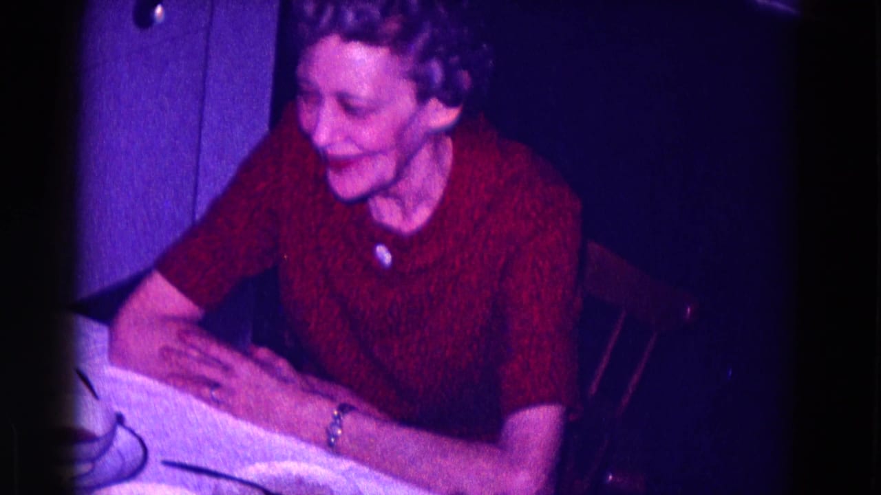 In 1968 Minnesota home-movie footage, an older woman in a red textured dress smiles while seated at a table in a dim interior.