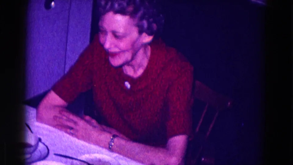 In 1968 Minnesota home-movie footage, an older woman in a red textured dress smiles while seated at a table in a dim interior.