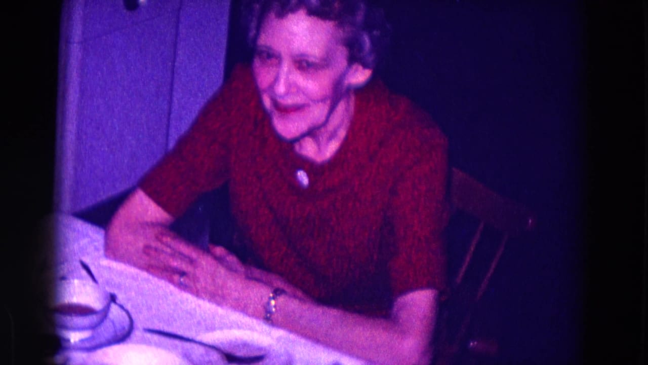 Older woman in a red dress looks toward the camera while seated beside a table setting.