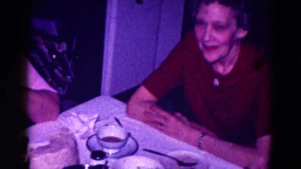 An older woman in a red dress sits by a white tablecloth and teacup, with another seated adult partly visible in the frame.